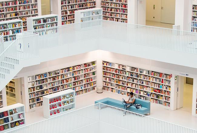 Multilevel library with many books, white floors and shelves, seating areas, and a staircase.