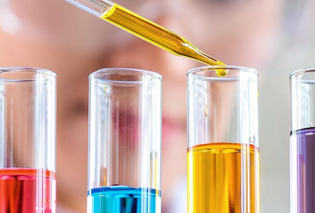 A woman is filling a liquid into a test tube using a pipette. Several test tubes with colorful liquids are in front of her.